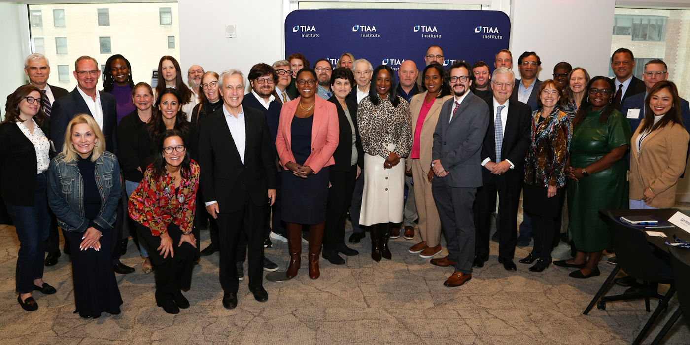A large group of approximately 35 professionals posing together for a group photo in front of a TIAA Institute branded step-and-repeat banner at the 2025 Higher Education Fellows Symposium.
