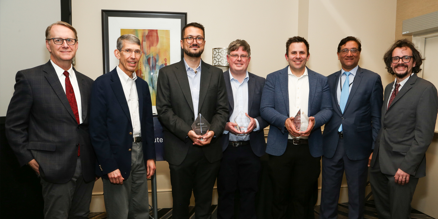 Seven professionals in business attire, three holding award trophies, pose for a group photo at the 2025 Samuelson Award ceremony.