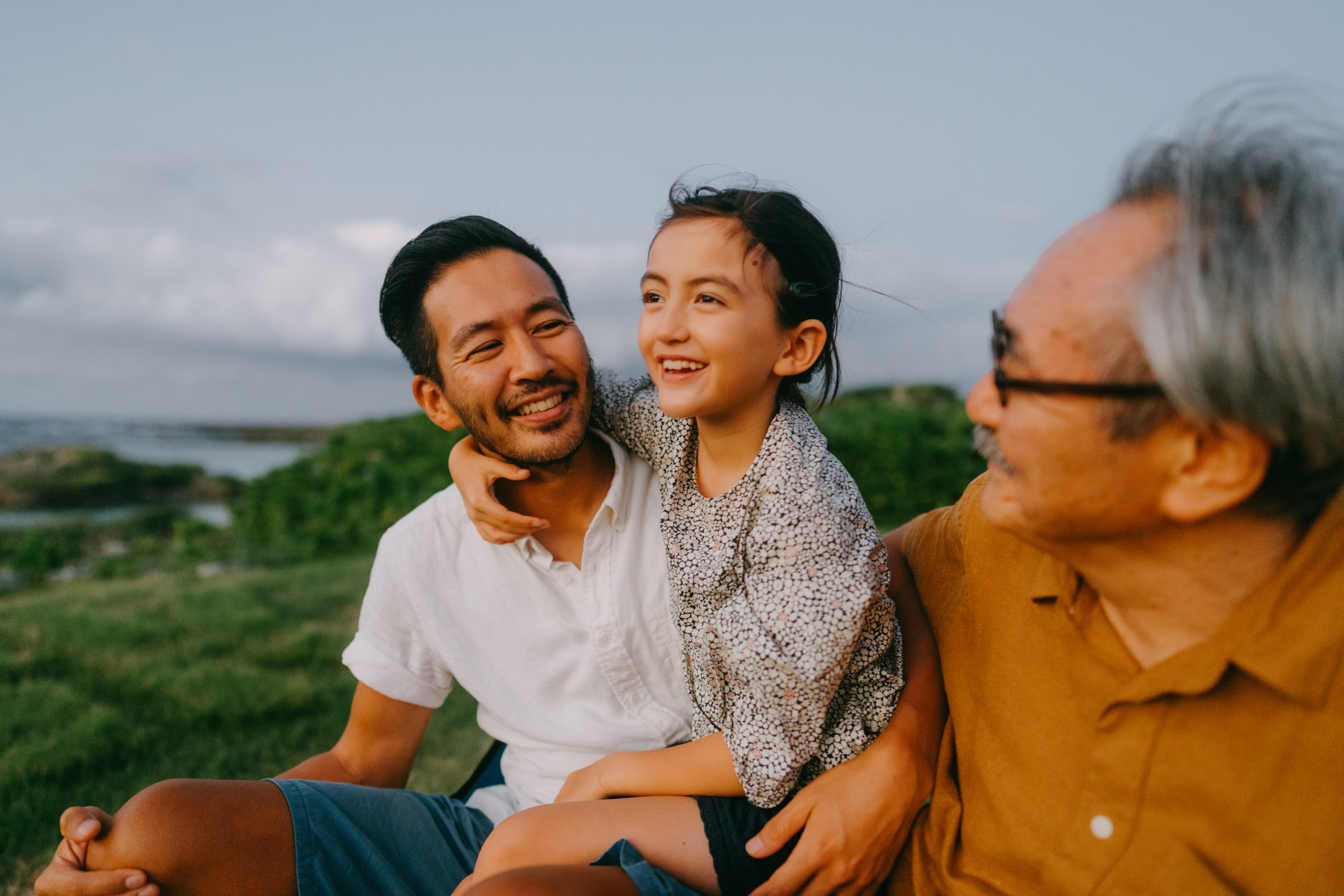 Click image of three generations sitting by seashore smiling; father and grandfather watch young girl as she gazes to sea to navigate to press release: A great place for working parents