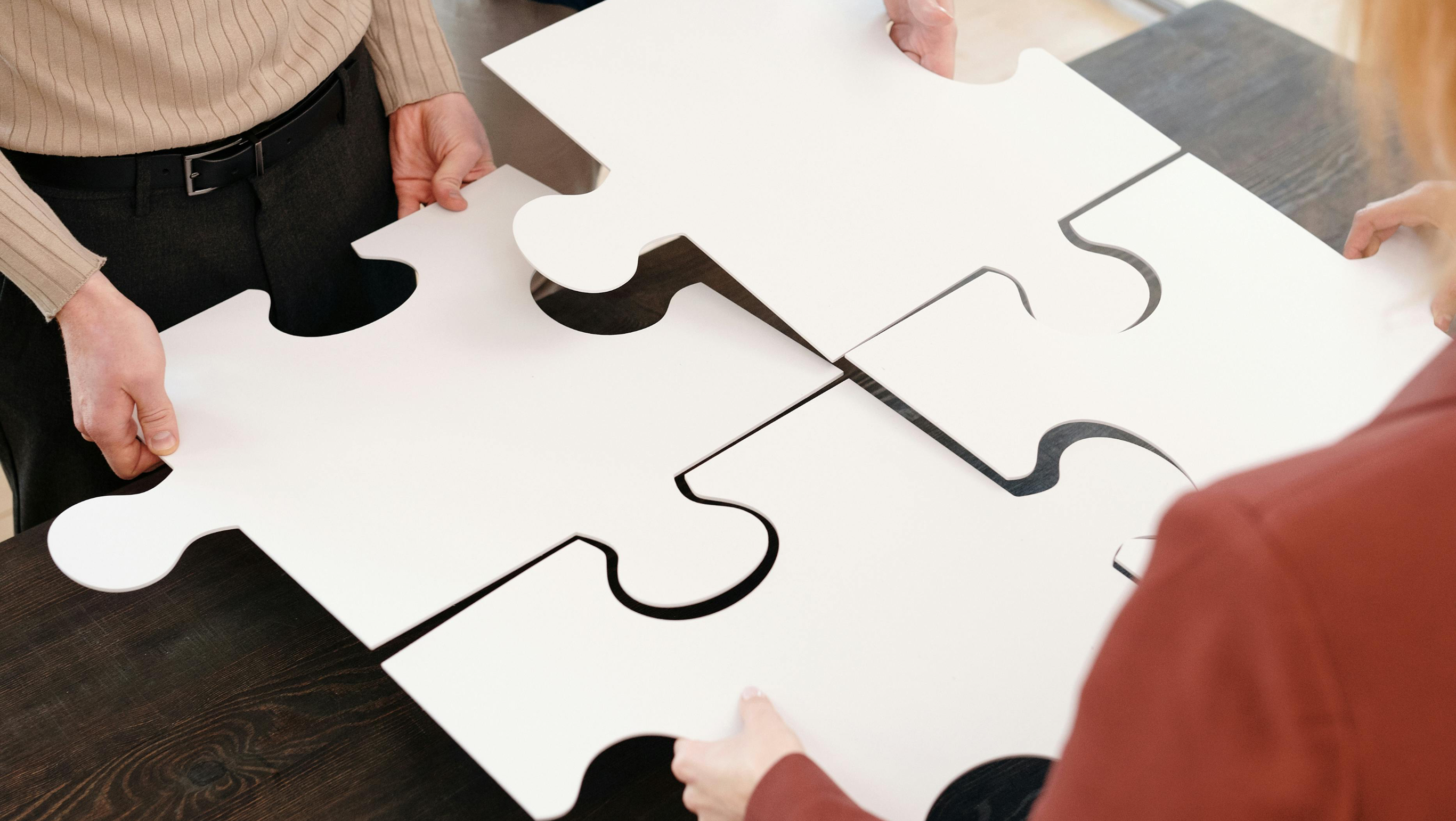Four professionals collaborate to fit together large white puzzle pieces on a wooden table, symbolizing teamwork and problem-solving.