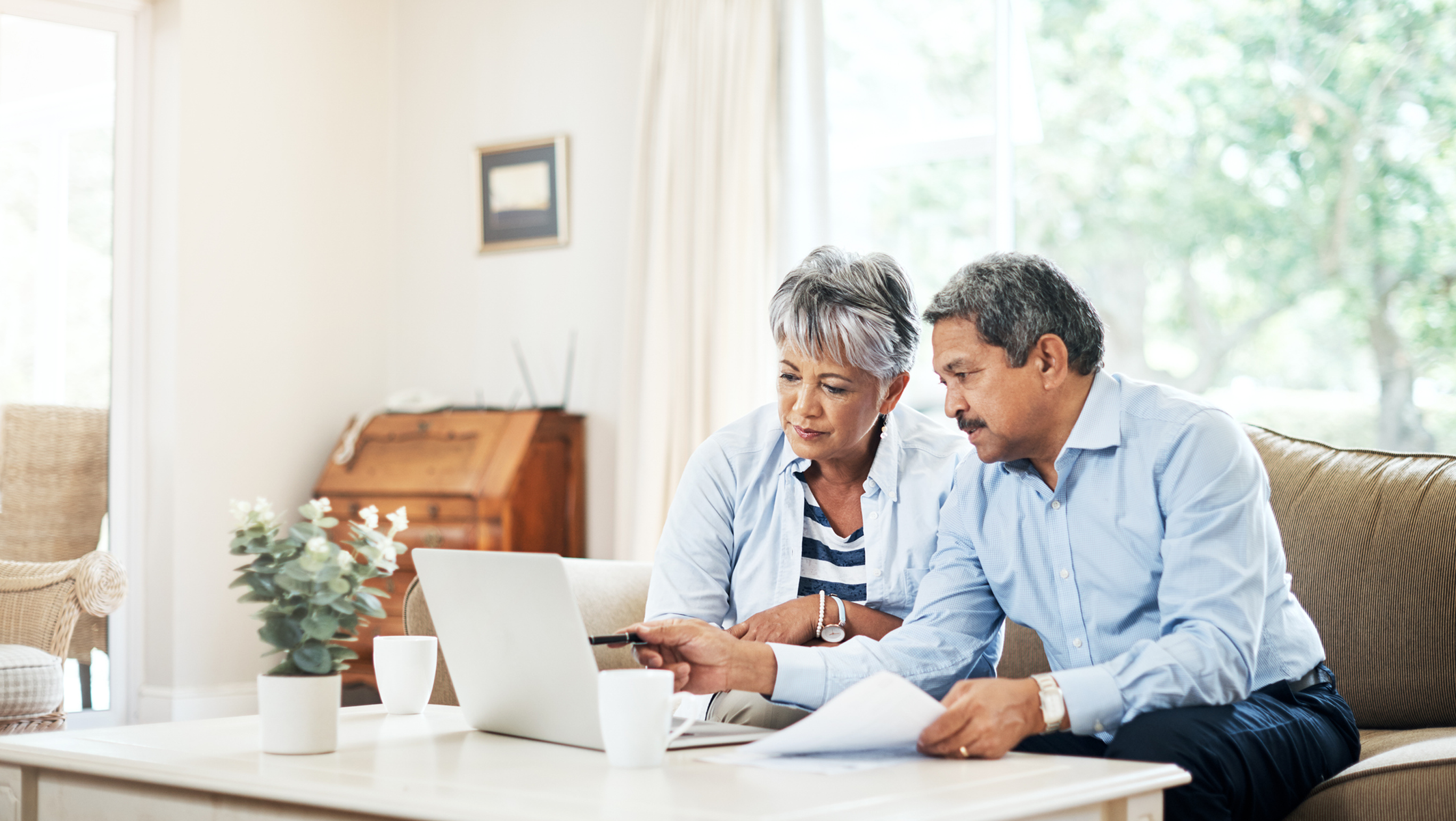 A couple in their 50s sit in their living room working on a laptop with papers spread around them.