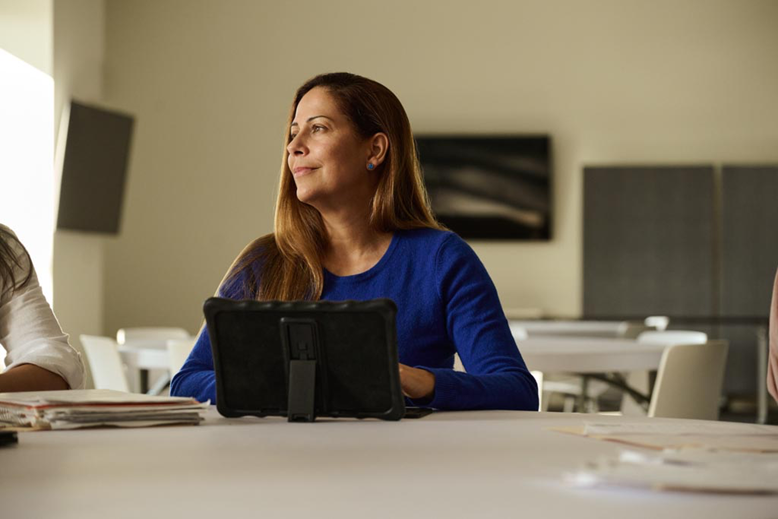 A woman with long brown hair wearing a blue sweater sits at a table with a tablet in front of her.