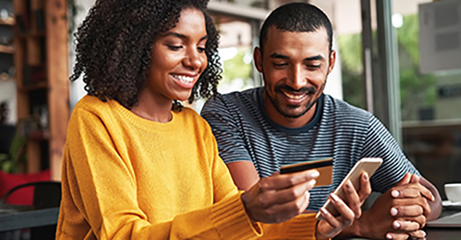 Young african man looking at his girlfriend using credit card and smartphone for shopping online in cafe