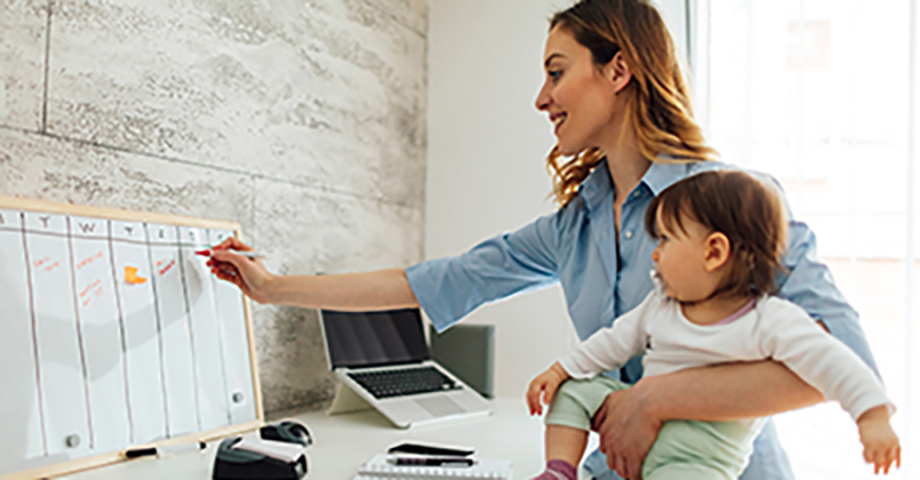 Mother working from home while carrying her cute baby girl. She is writing on whiteboard, entering into the schedule. She is happy and cheerful.